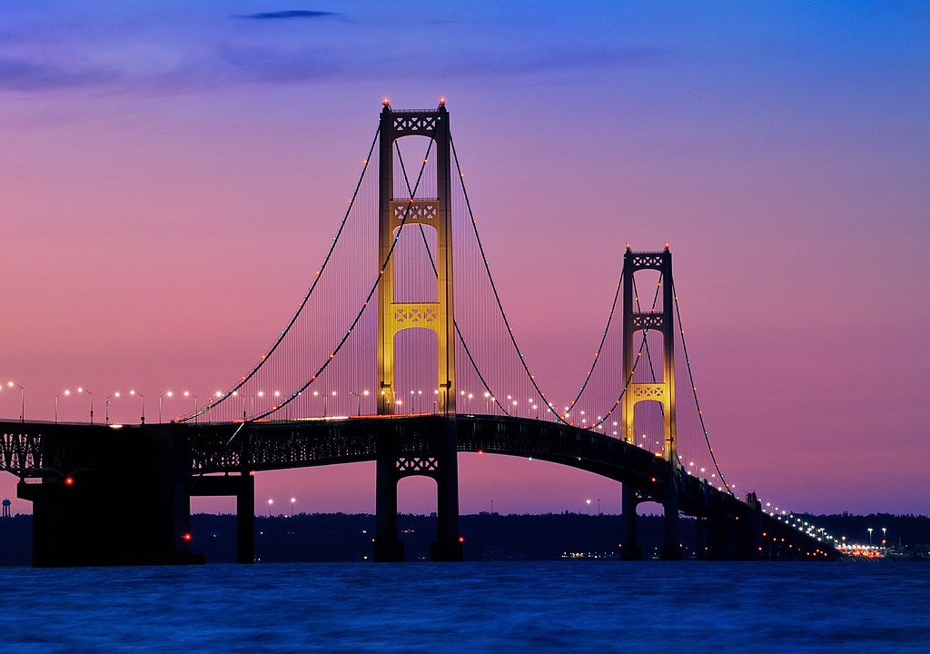 The Mackinac Bridge lit up at dusk, its towers glowing gold against a violet sky
